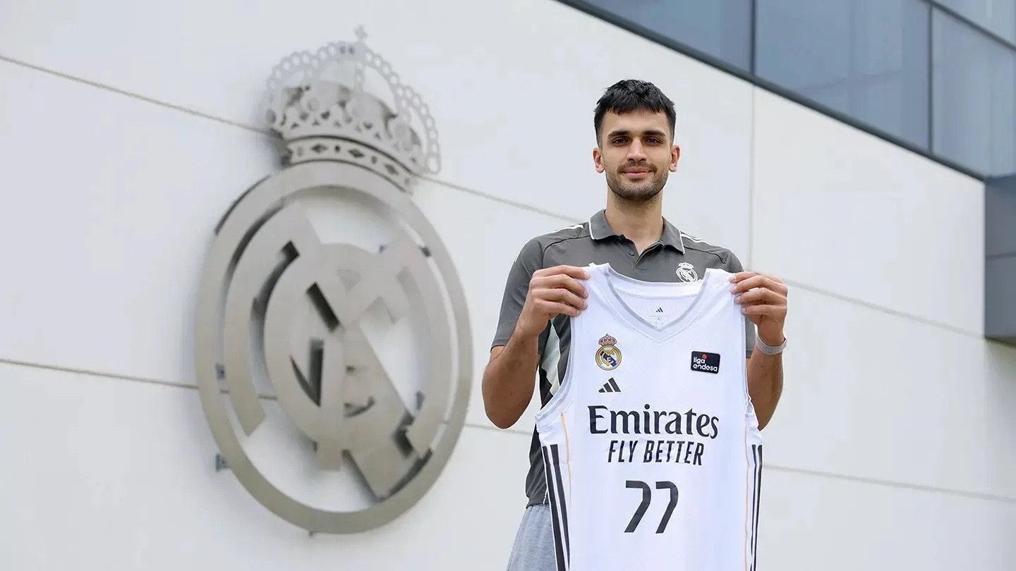Ömer Yurtseven posa con la camiseta del Real Madrid junto al pabellón de baloncesto de la Ciudad Real Madrid, donde se ve el escudo del equipo detrás de él.